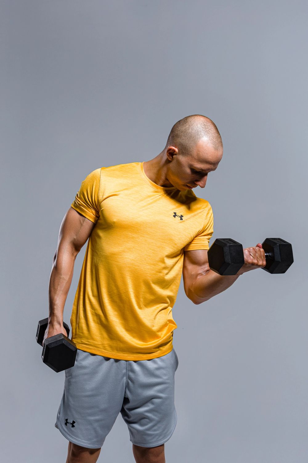 Studio fitness portret muškarca tokom vežbe sa bučicama, snimljen na neutralnoj pozadini, sa naglaskom na koncentraciju, kontrolu pokreta i mišićnu definiciju ruku, uz čisto i kontrolisano osvetljenje.