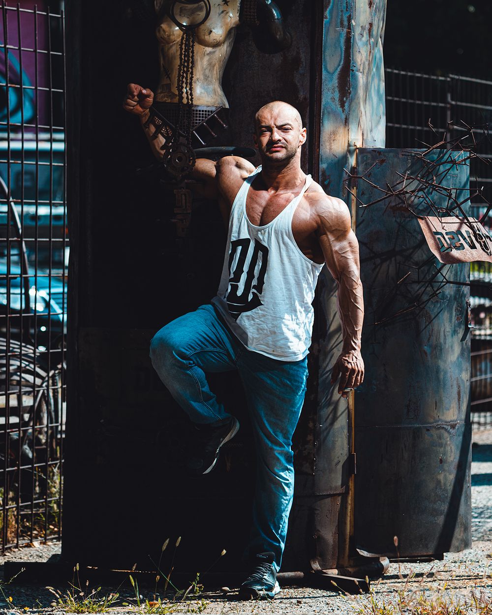 Urban fitness portret muškarca u industrijskom okruženju, sa naglašenom mišićnom definicijom i snažnim stavom, snimljen uz metalne strukture, sa kontrastnim osvetljenjem i fokusom na snagu, izdržljivost i karakter.