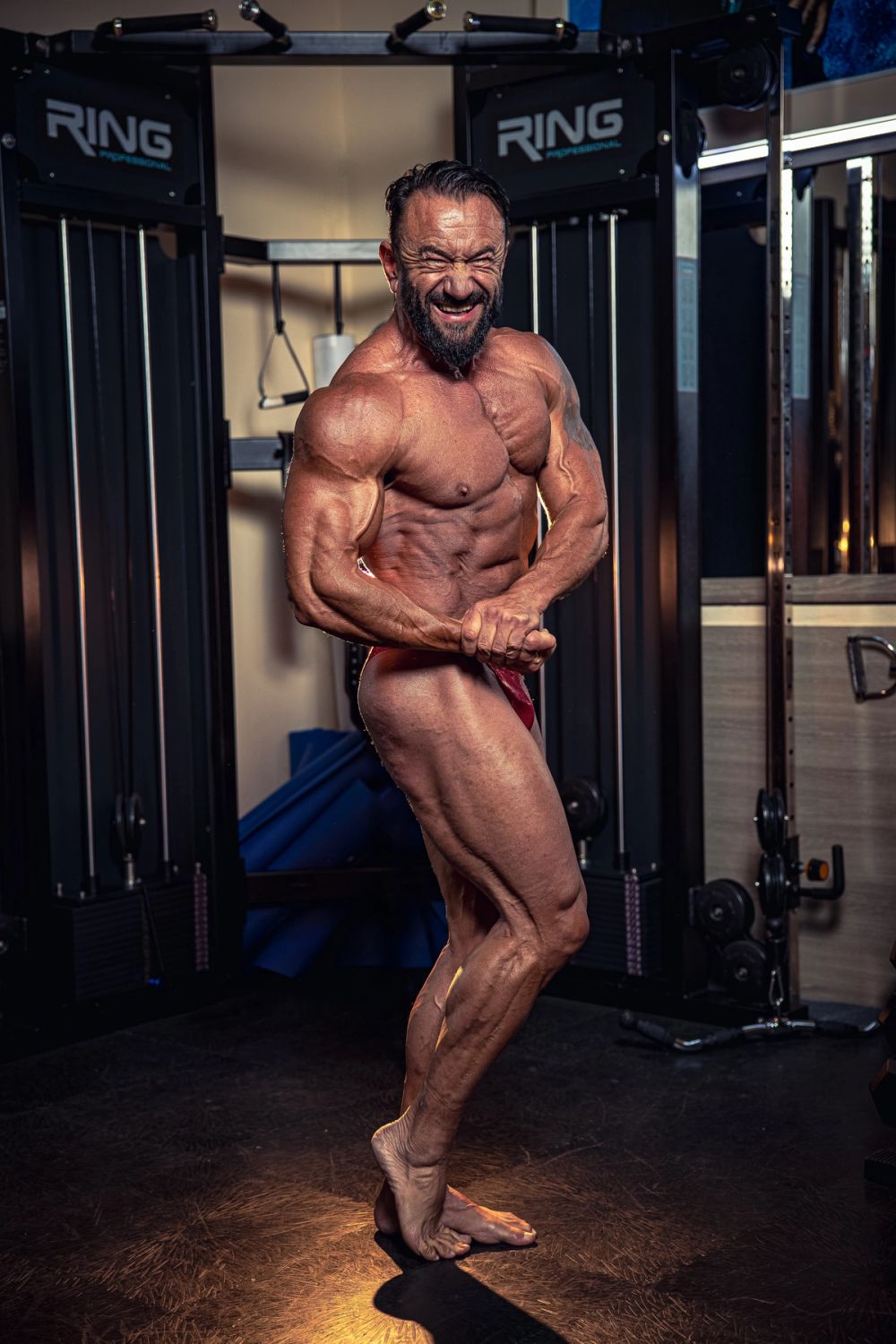 Profesionalni bodybuilding portret muškarca u teretani tokom poziranja, sa izrazitom mišićnom definicijom celog tela, naglašenim kontrakcijama i dramatičnim osvetljenjem, sa fokusom na vrhunsku fizičku formu.