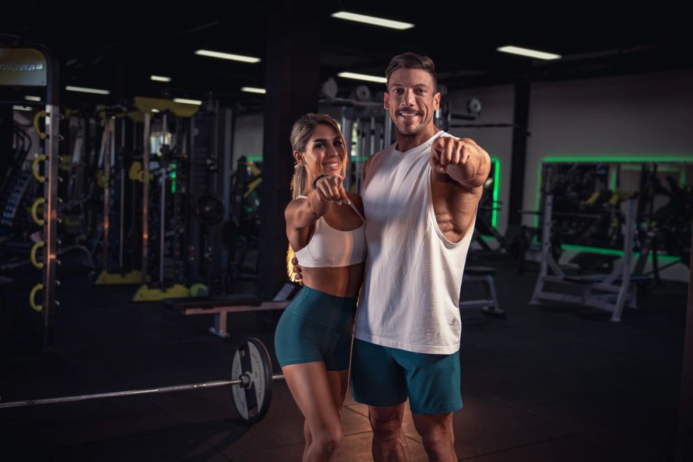Fitness portret muškarca i žene u teretani, u sportskom outfitu, kako stoje zajedno i pokazuju ka kameri, sa naglaskom na energiju, timski duh i motivaciju za trening.
