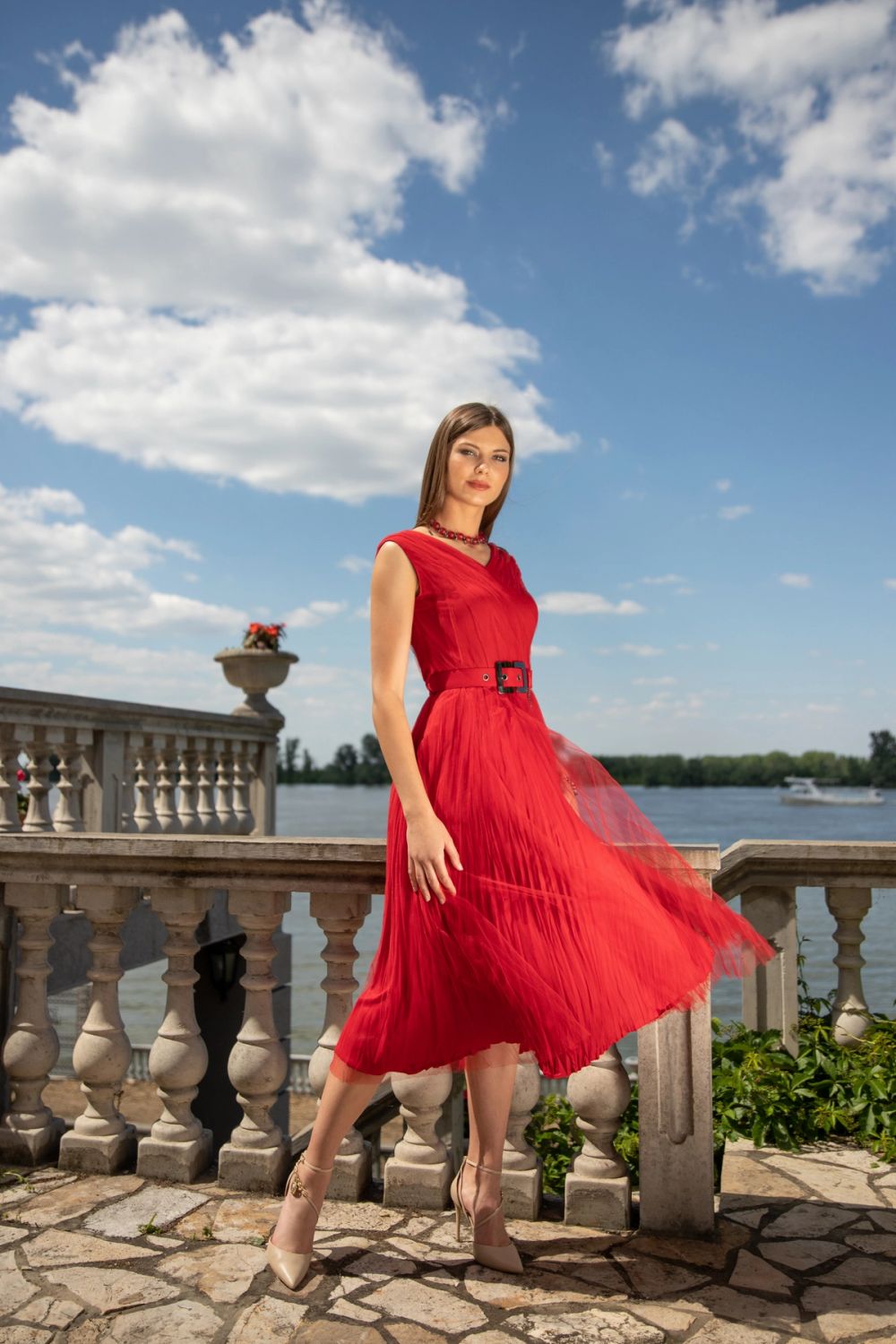 Model u elegantnoj crvenoj haljini pozira na otvorenom, fotografisana u prirodnom svetlu, sa naglaskom na pokret tkanine i lifestyle modni stil.