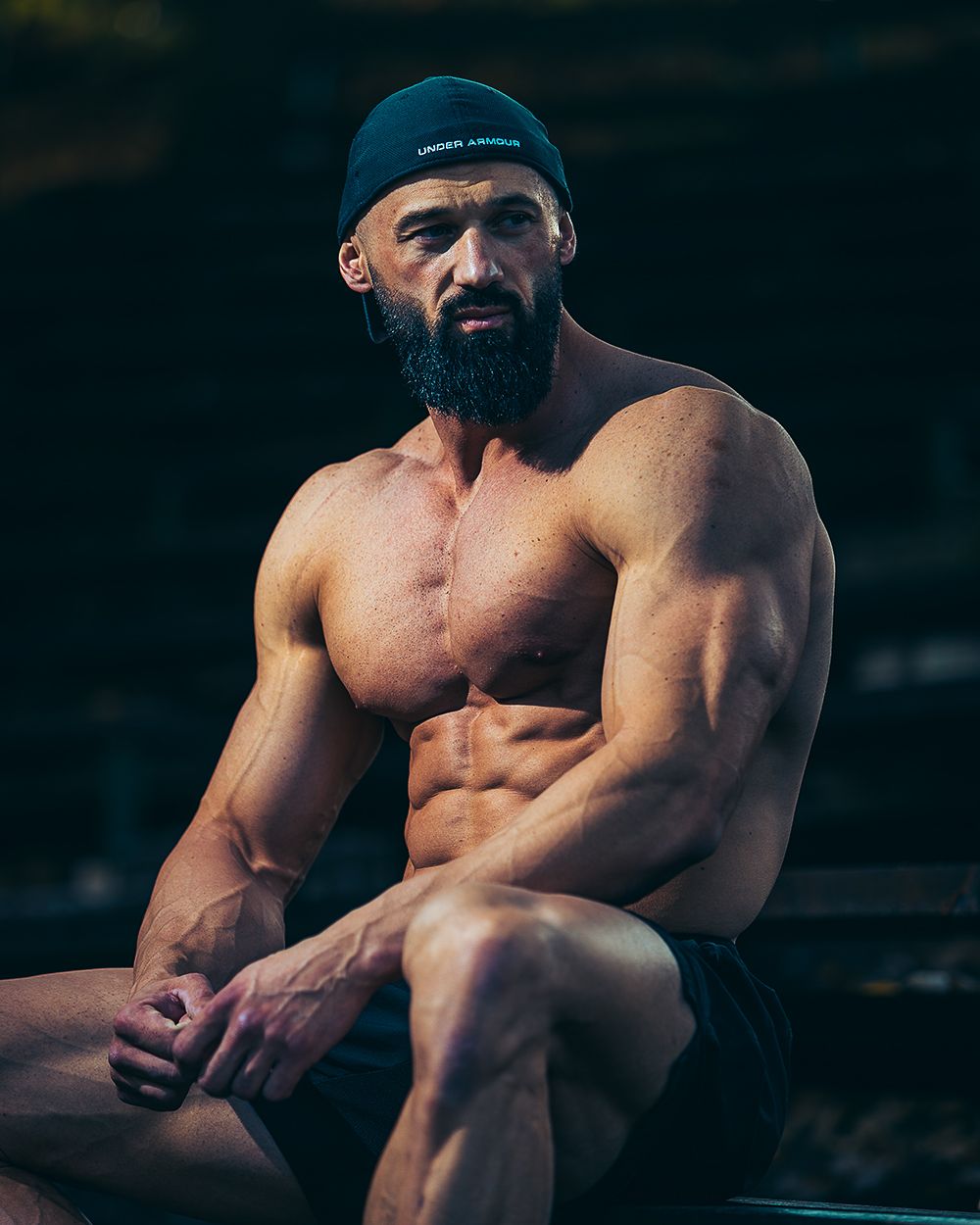 Fitness portret muškarca u teretani tokom pauze na treningu, sa izraženom mišićnom definicijom, snažnim kontrastnim osvetljenjem i fokusom na koncentraciju i fizičku spremu.