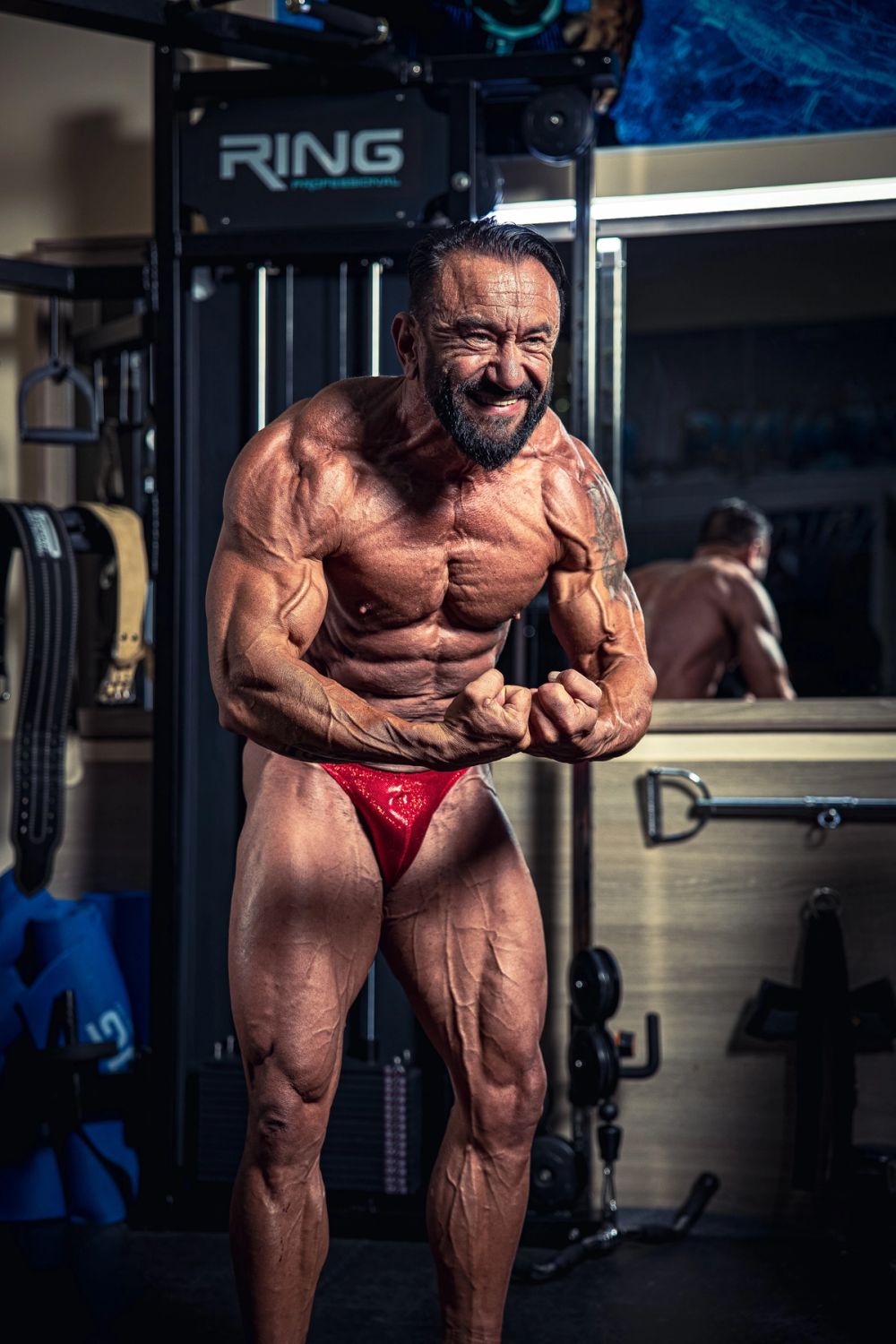 Profesionalni bodybuilding portret muškarca u teretani tokom intenzivnog poziranja, sa izrazitom mišićnom definicijom celog tela, snažnim kontrakcijama i dramatičnim osvetljenjem, uz fokus na vrhunsku fizičku formu i snagu.
