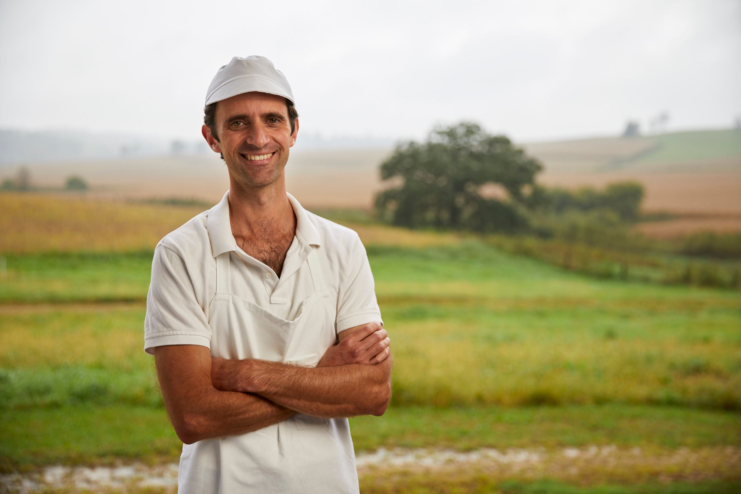 A smiling cheesemaker in a white cap with arms crossed, standing in a misty pasture at Uplands Cheese
