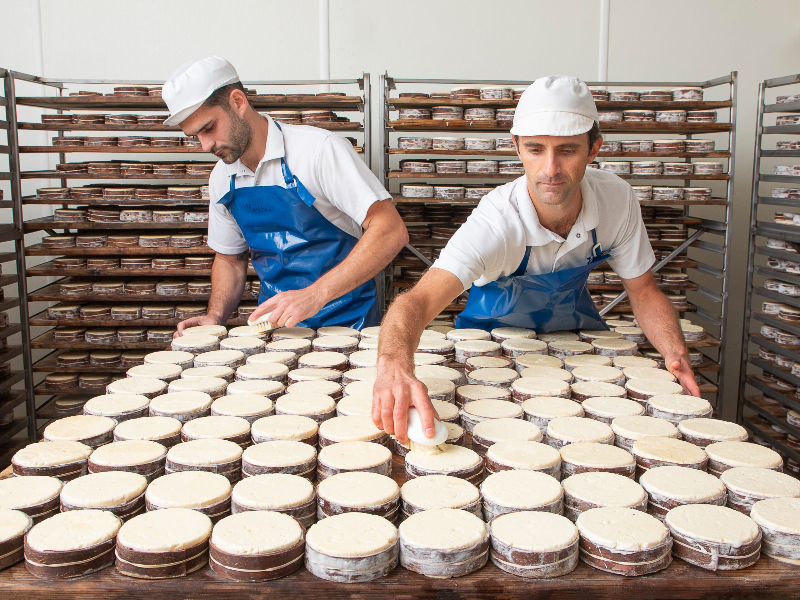 Two cheesemakers washing rows of bark-banded Rush Creek Reserve wheels in the cave, with aging racks behind them