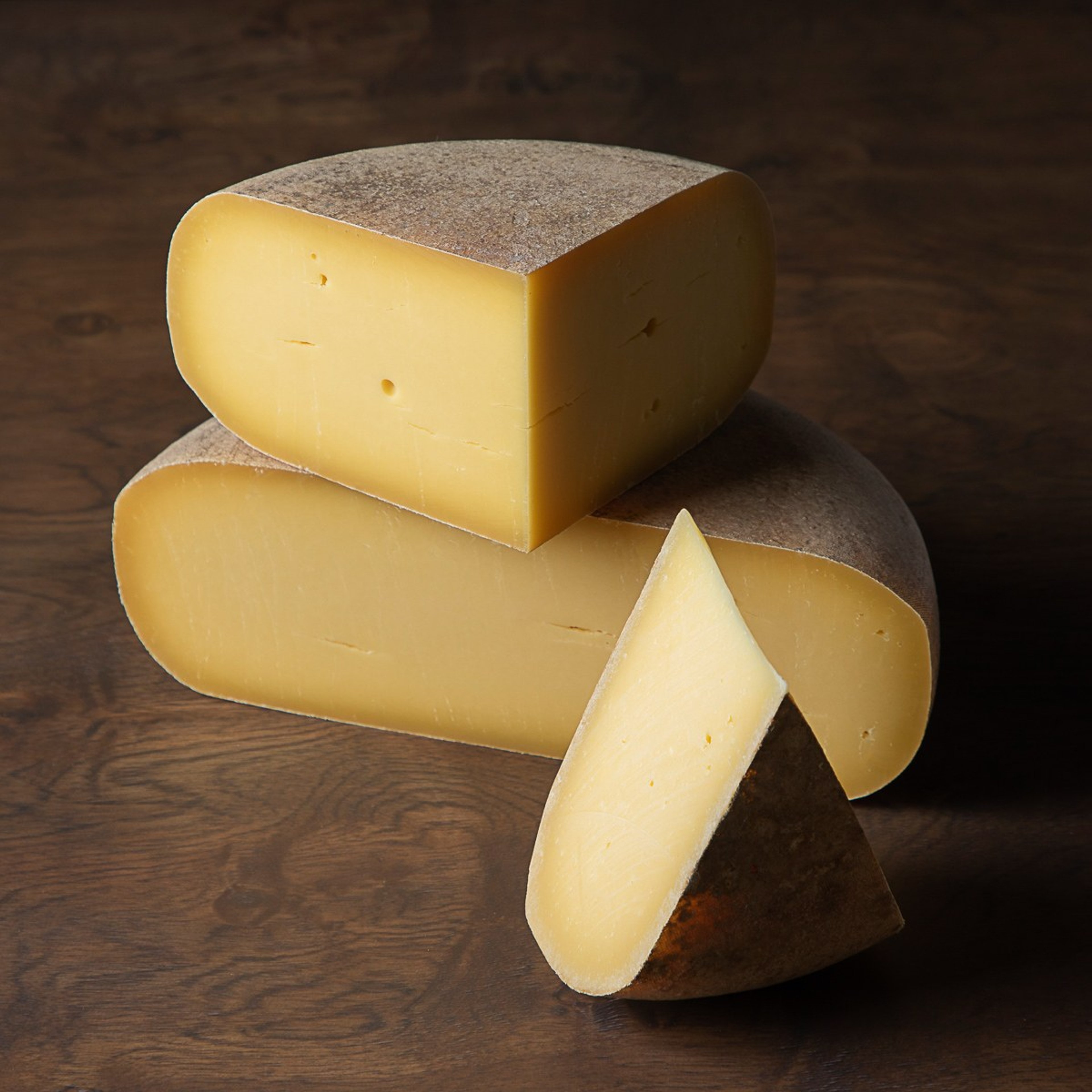 A half wheel and a wedge of Pleasant Ridge Reserve stacked on a dark wooden table