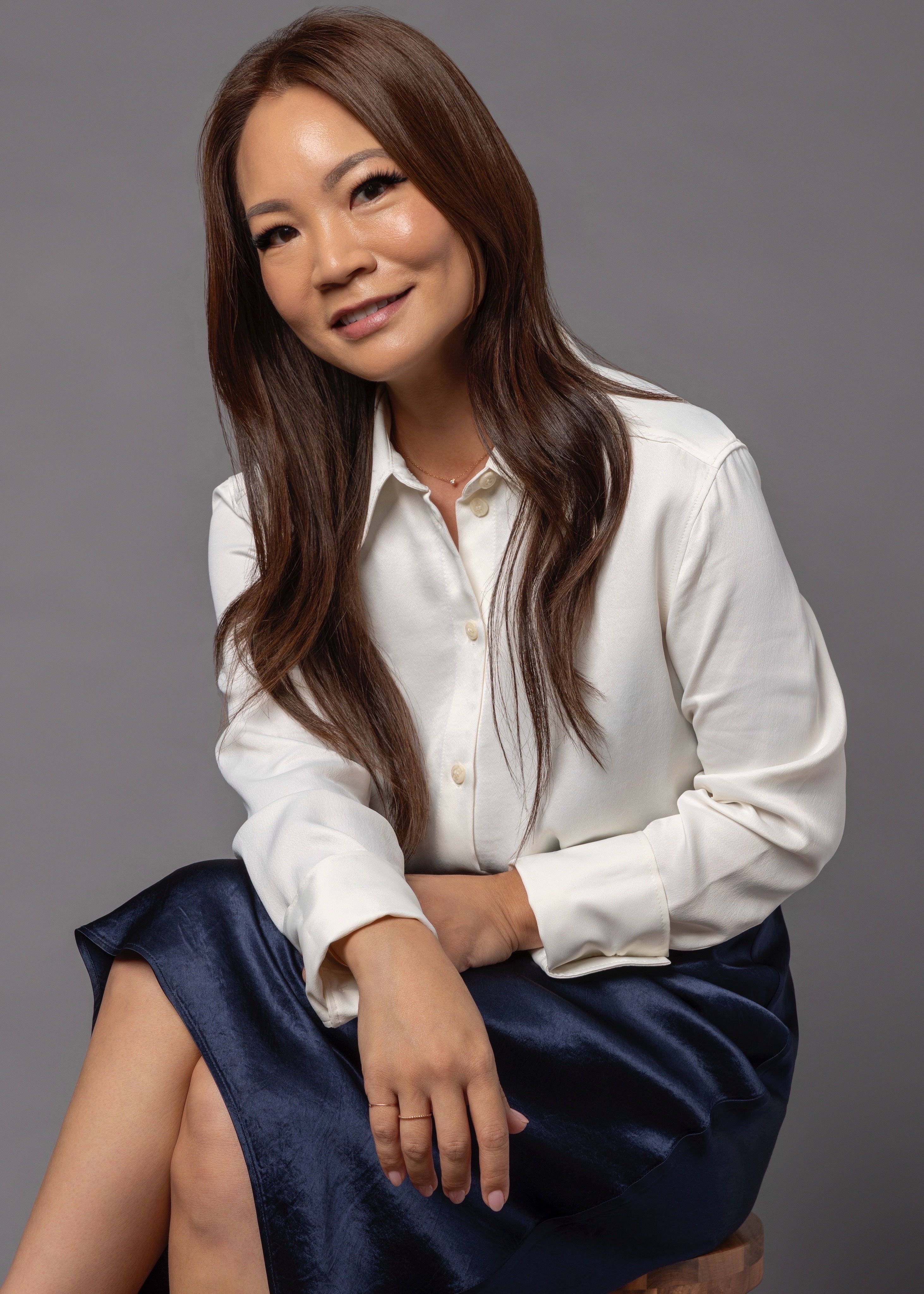 A smiling woman with long brown hair, wearing a cream shirt and navy satin skirt, sits against a grey background.