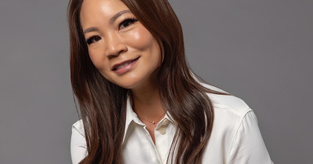 A smiling woman with long brown hair, wearing a cream shirt and navy satin skirt, sits against a grey background.
