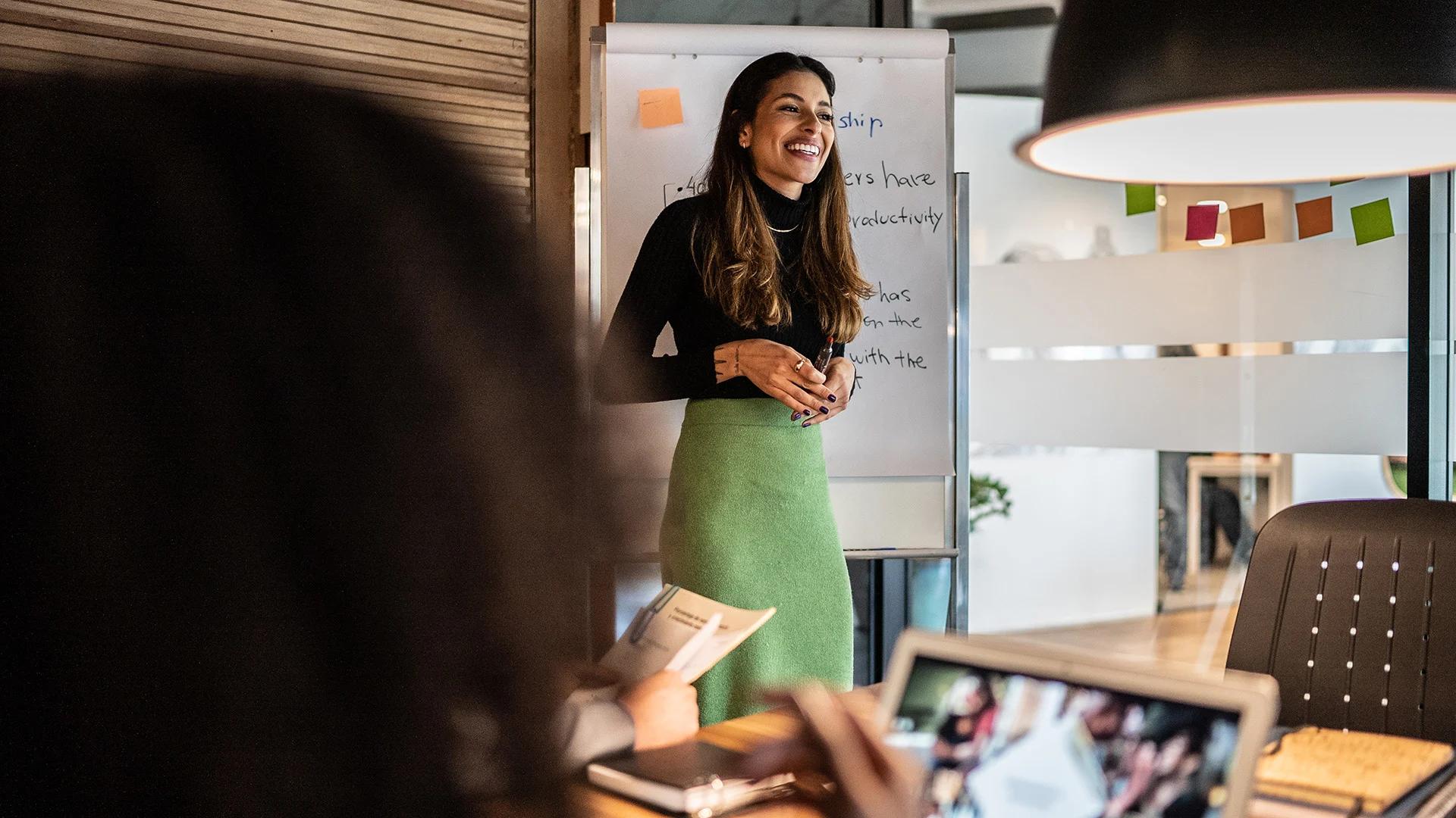 A smiling woman presents in front of a whiteboard to a group in an office meeting.