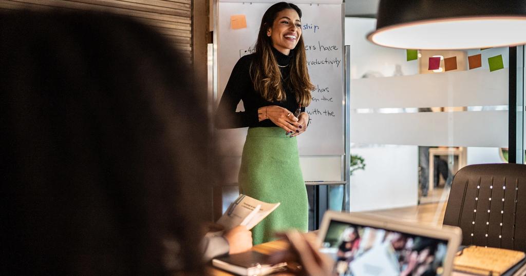 A smiling woman presents in front of a whiteboard to a group in an office meeting.