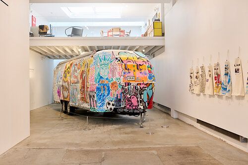 An Airstream trailer covered in colorful graffiti art is displayed in a modern gallery, with decorated aprons hanging on a white wall.