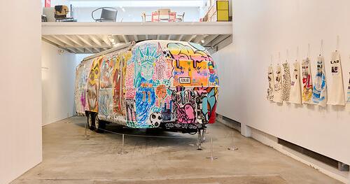 An Airstream trailer covered in colorful graffiti art is displayed in a modern gallery, with decorated aprons hanging on a white wall.