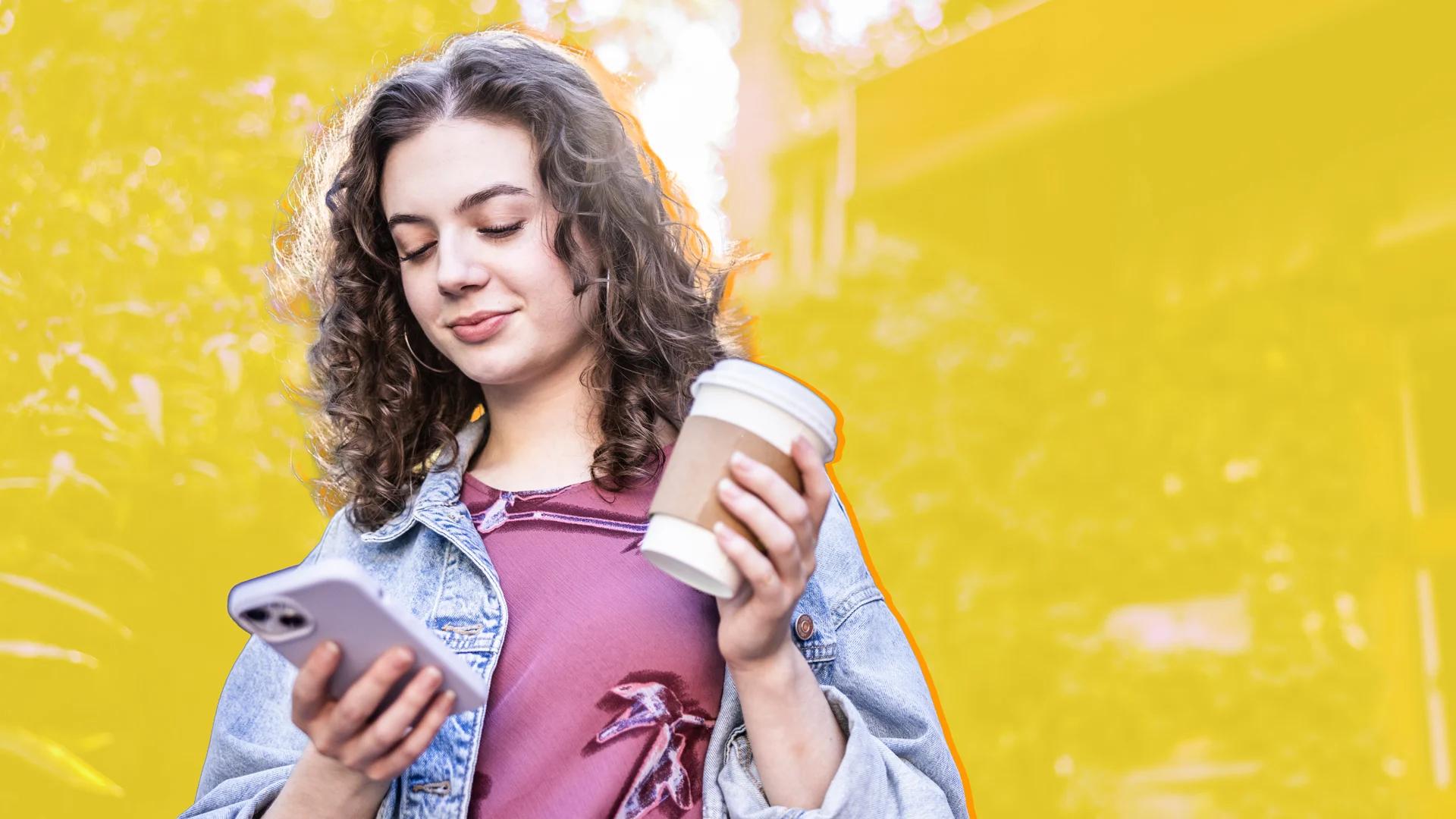 A young woman with curly hair smiles while looking at her phone and holding a coffee cup.