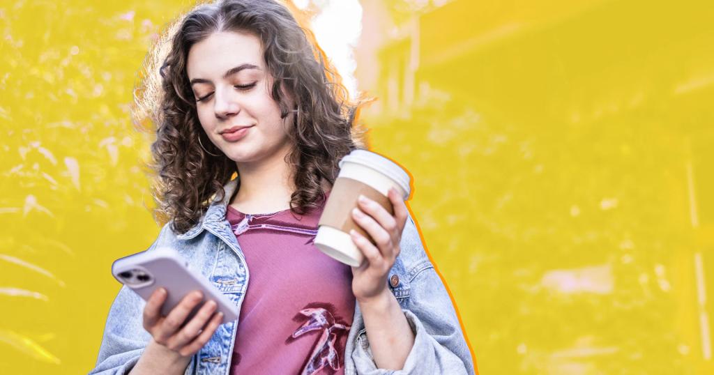 A young woman with curly hair smiles while looking at her phone and holding a coffee cup.