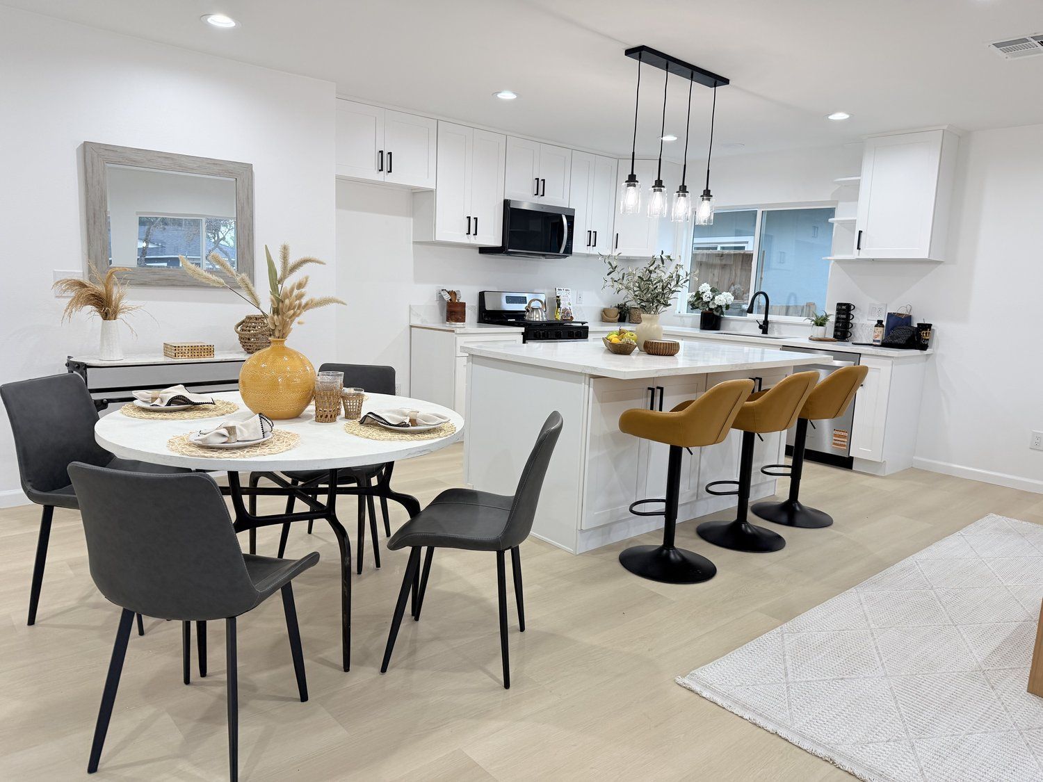 A contemporary dining area with a round marble table, black chairs, and a yellow vase, opening to a bright kitchen