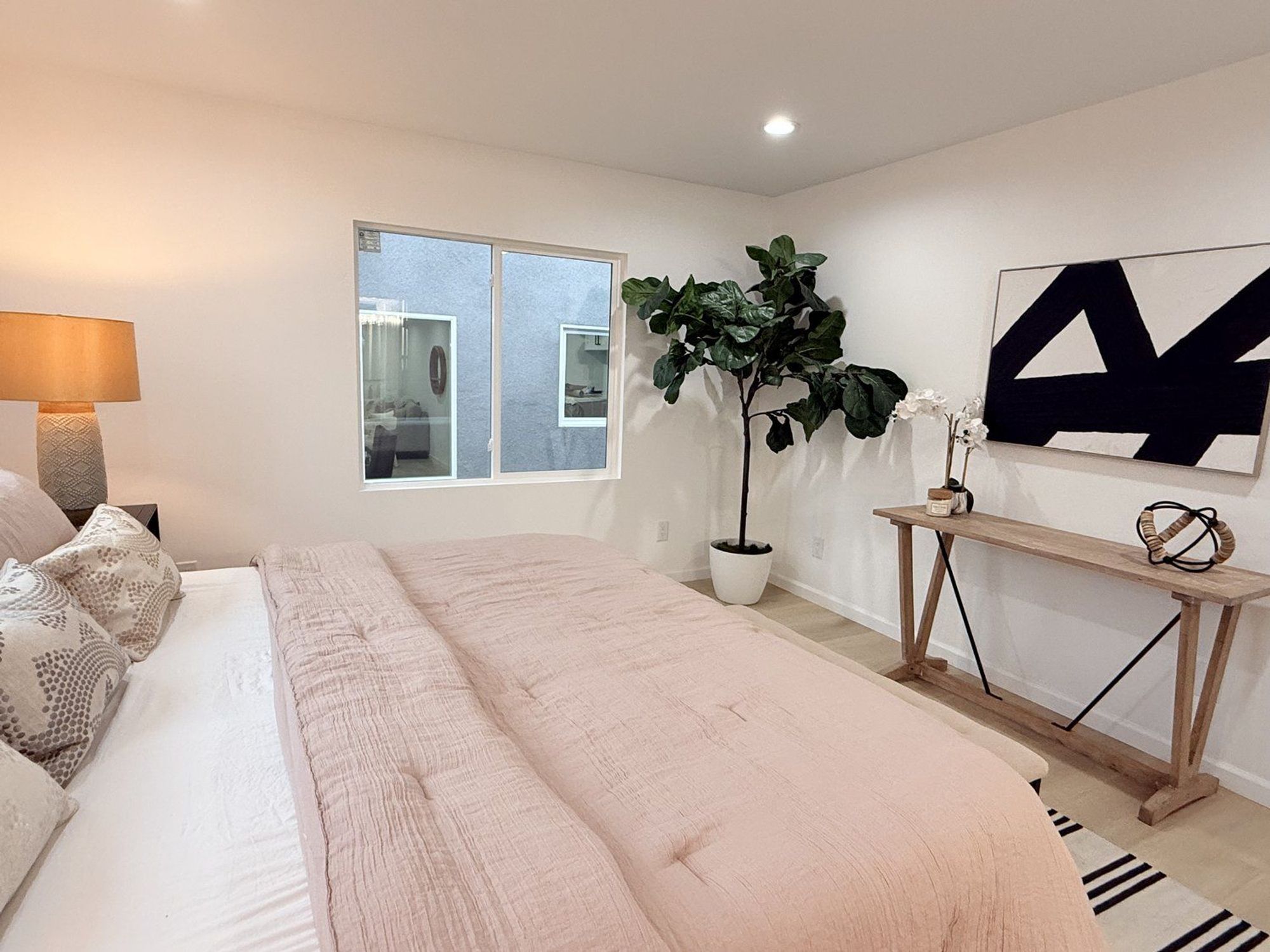 A bedroom with a light pink bedspread, wooden console table, potted plant, and black and white art