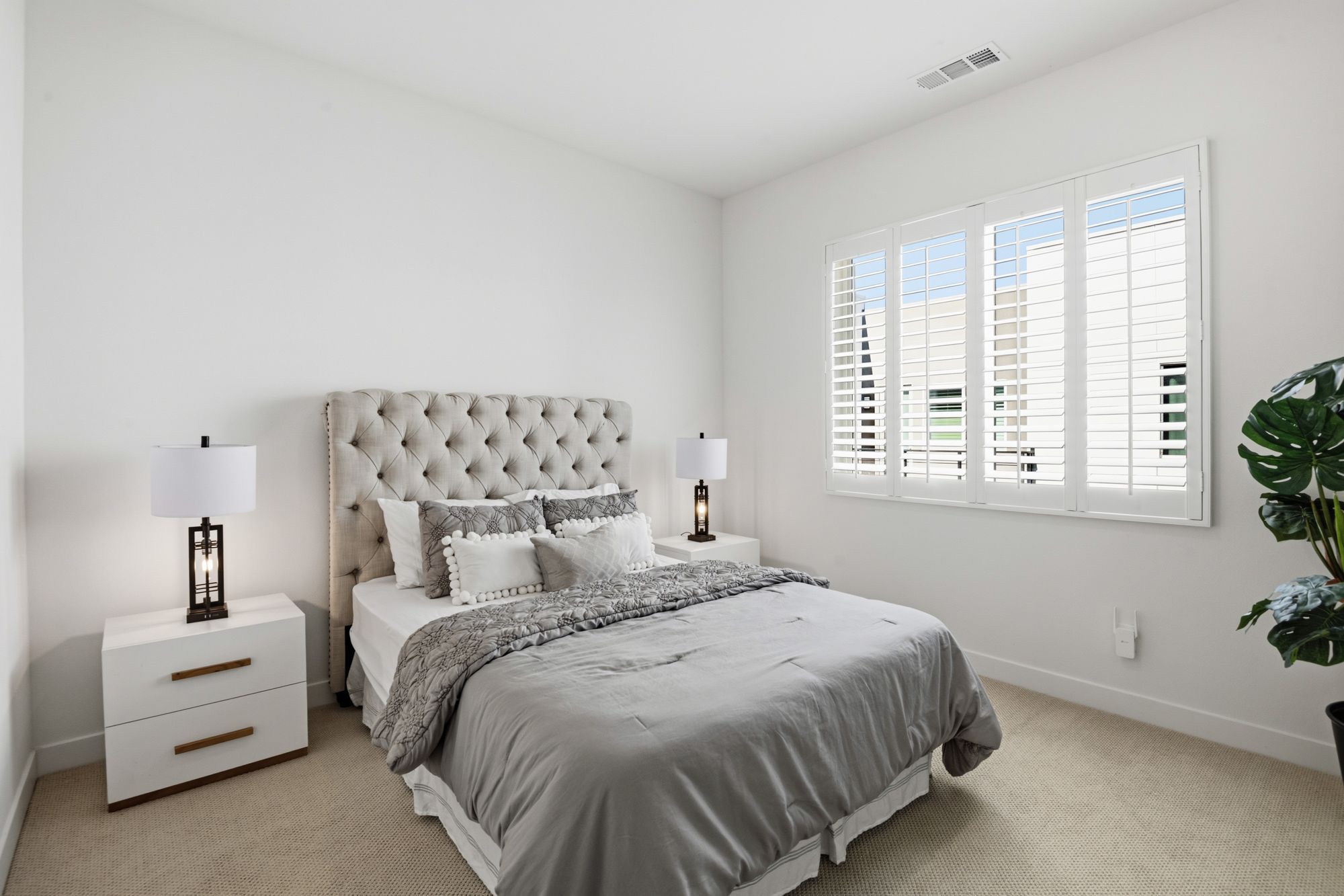 A modern bedroom with a tufted headboard, grey bedding, and a window with shutters