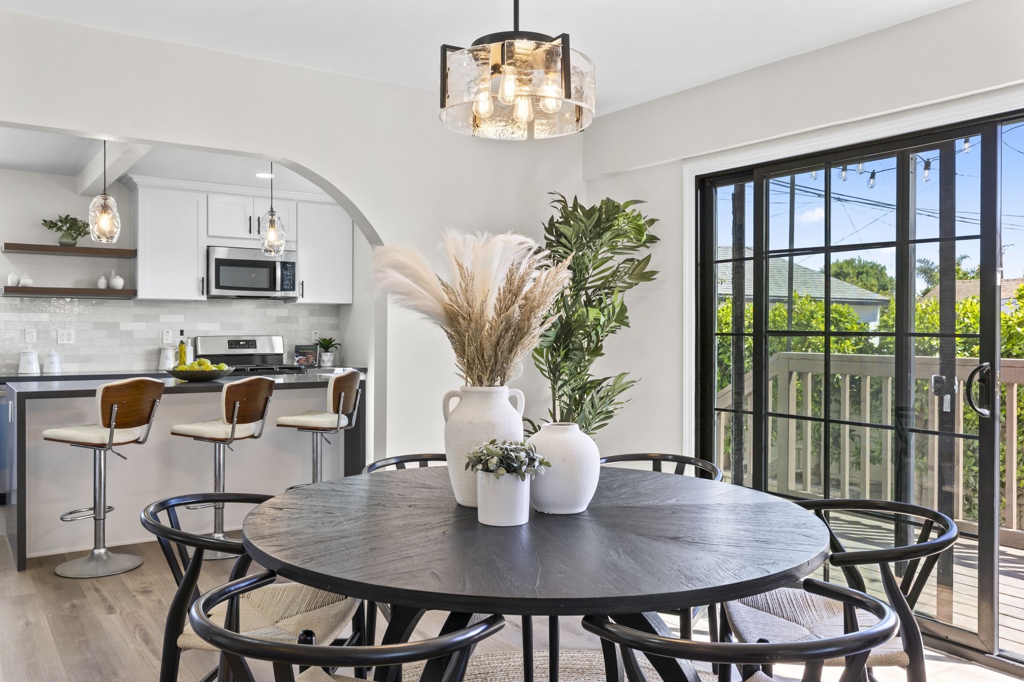 A modern dining area with a round black table, stylish chairs, and a vase of pampas grass, leading towards an open kitchen