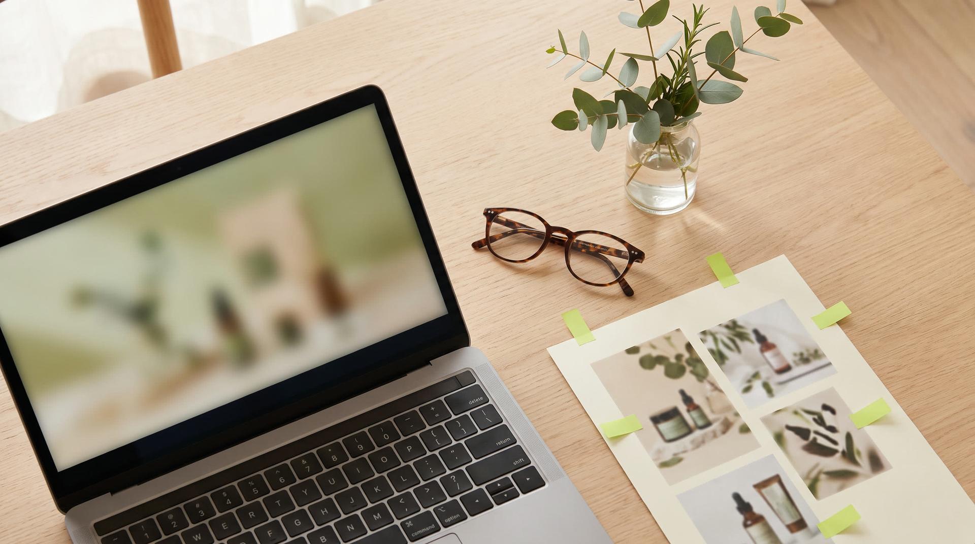Top-down view of a pale oak desk with a laptop, a printed sheet of product photos with green index tabs, reading glasses and a sprig of leaves in a glass