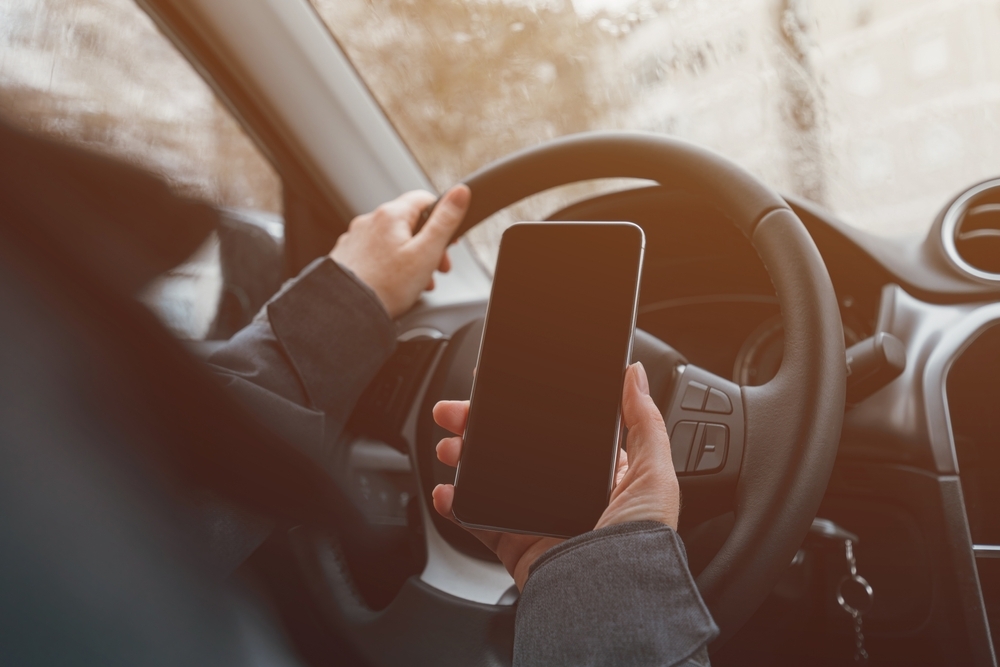 Image Driver holding a smartphone behind the steering wheel, illustrating distracted driving risk during the holiday shopping season.