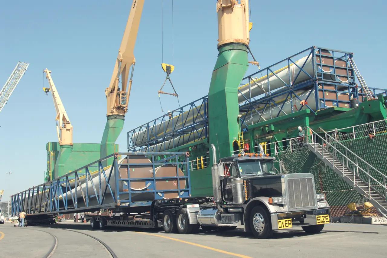 An oversize wind-turbine blade on a specialised trailer frame beside ship cranes