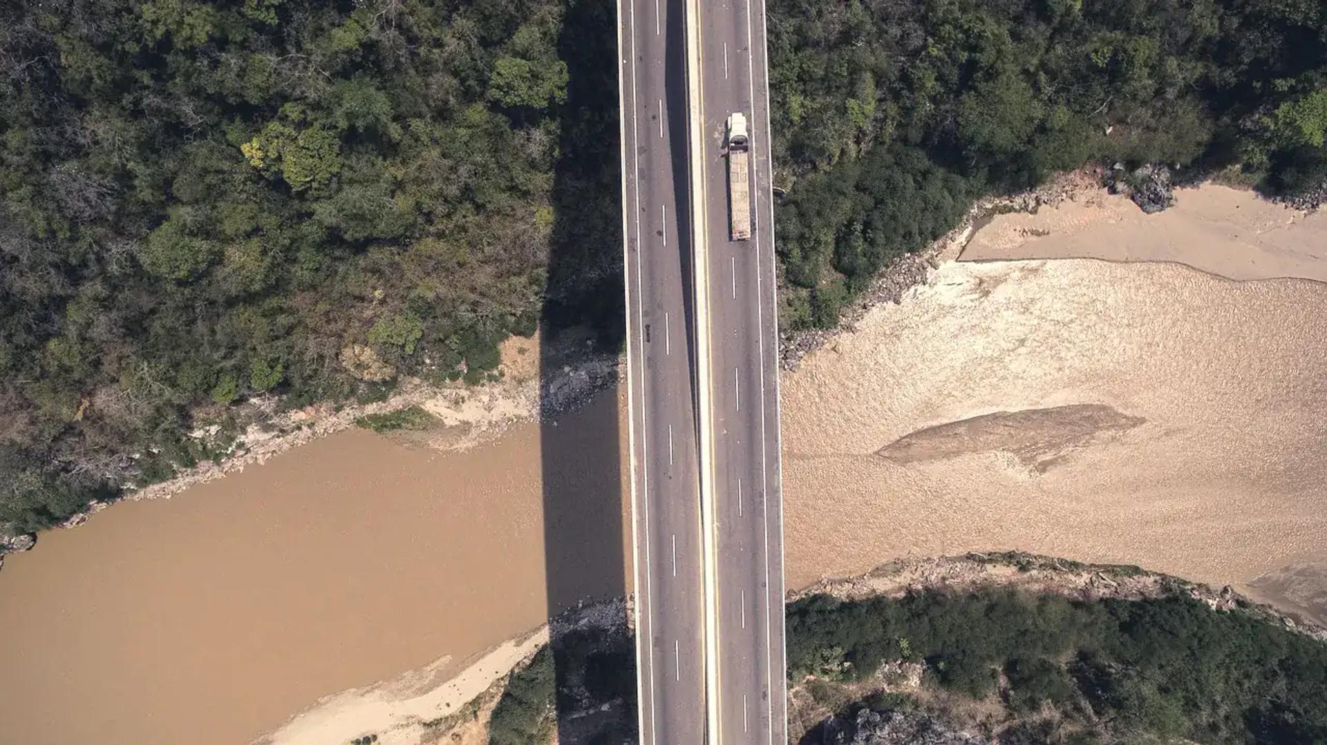 A lorry crossing a long road bridge over a brown river through dry bush, seen from above