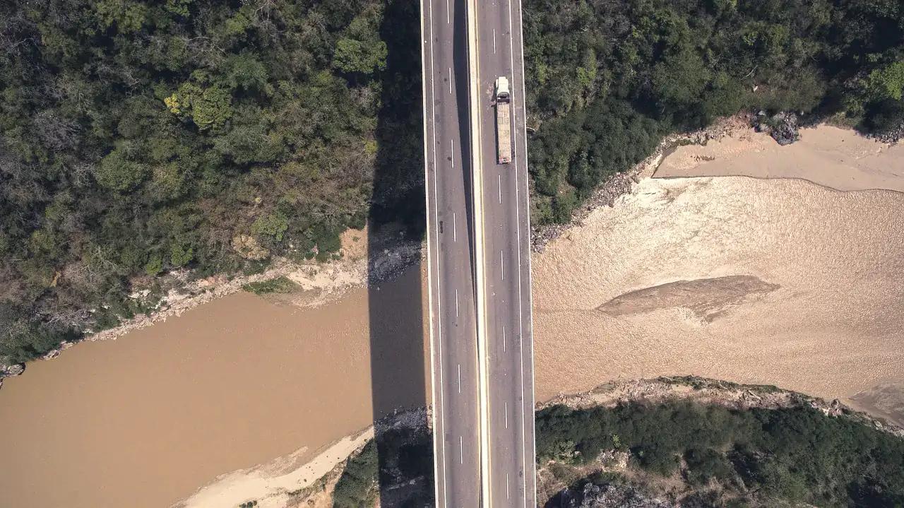 A lorry crossing a long road bridge over a brown river through dry bush, seen from above