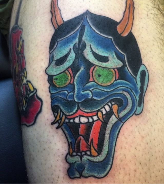 A colorful oni mask tattoo located on a person's thigh.