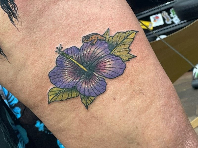 Tattoo of a frog on a hibiscus flower located on the upper arm.