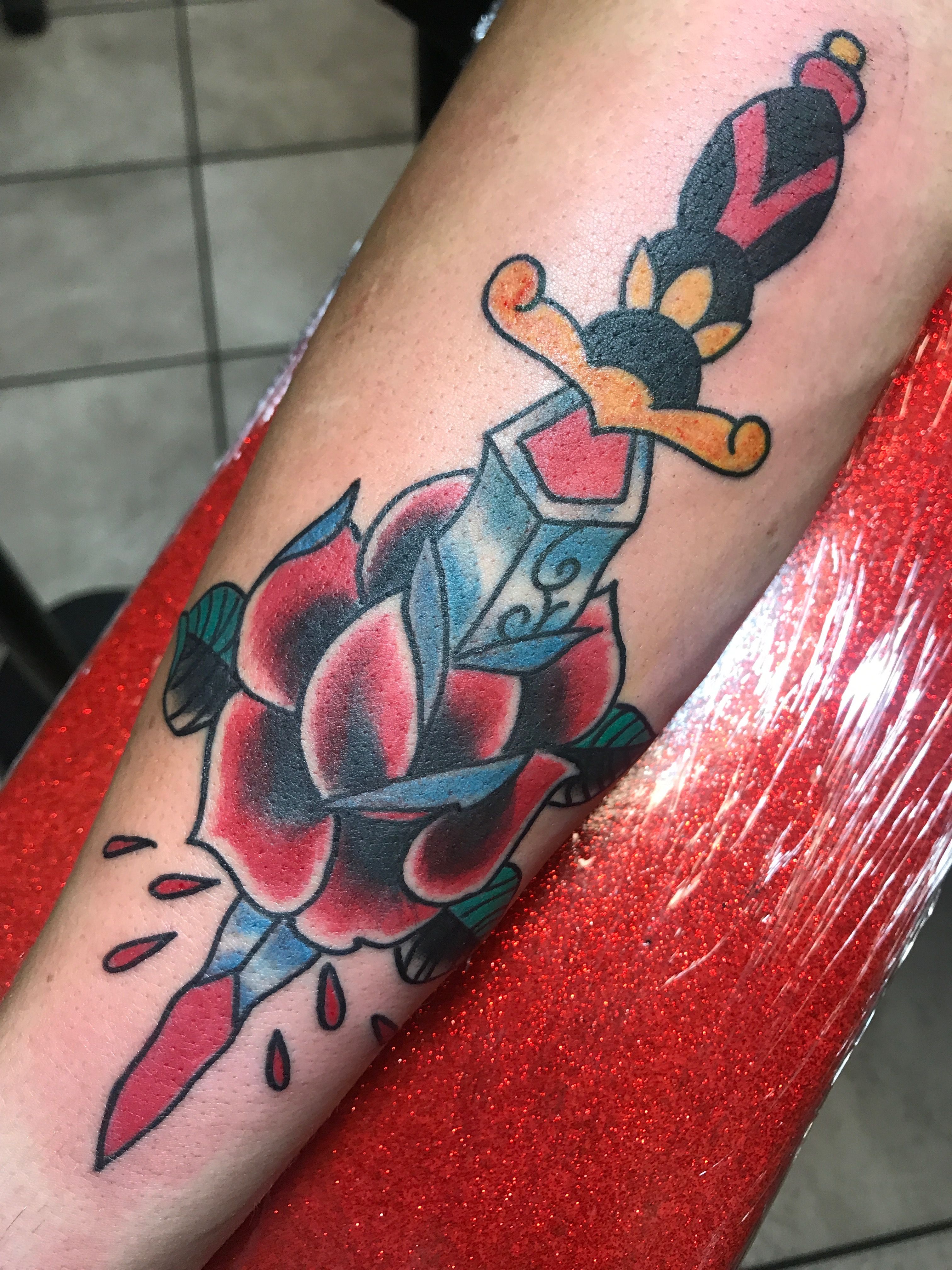 Tattoo of a dagger piercing a rose on a forearm with bold colors.