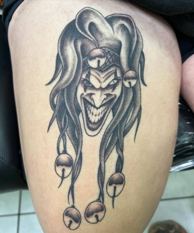 Tattoo of a menacing jester face in black and grey on a thigh.