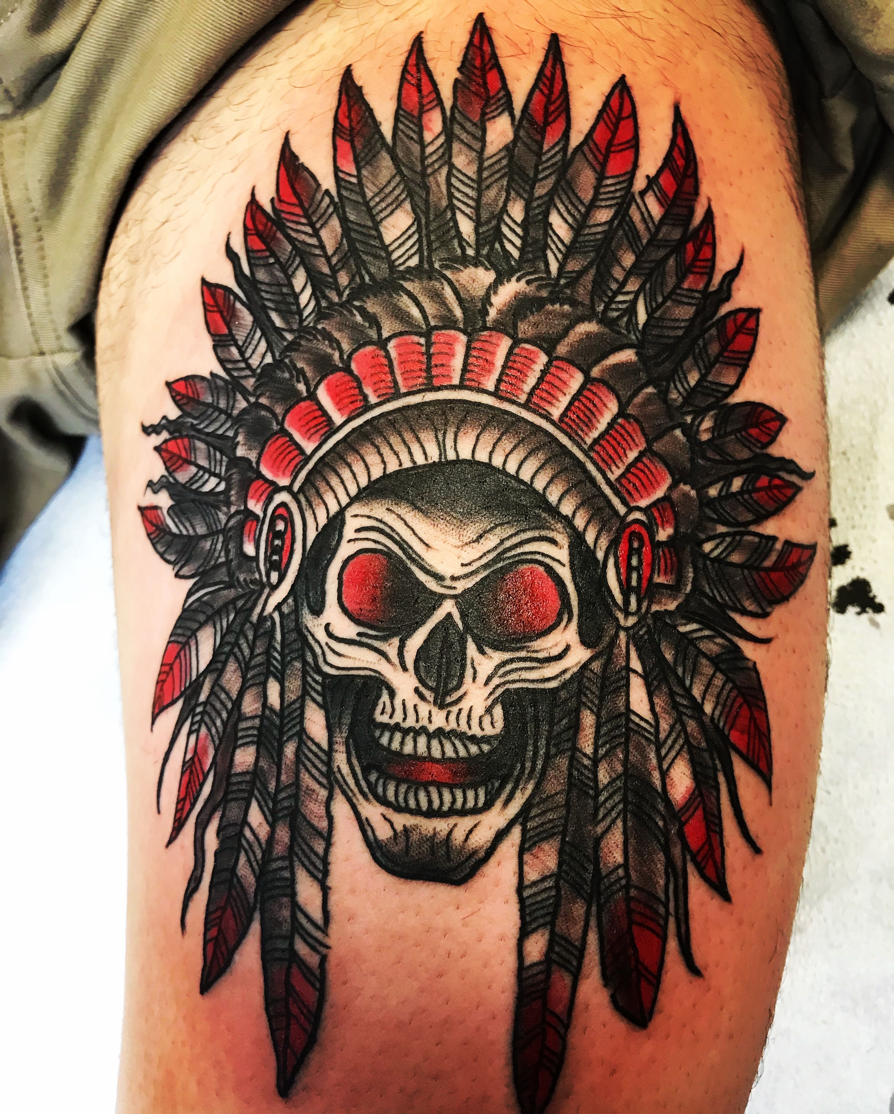 Tattoo of a skull wearing a feathered headdress in red and black on a thigh.