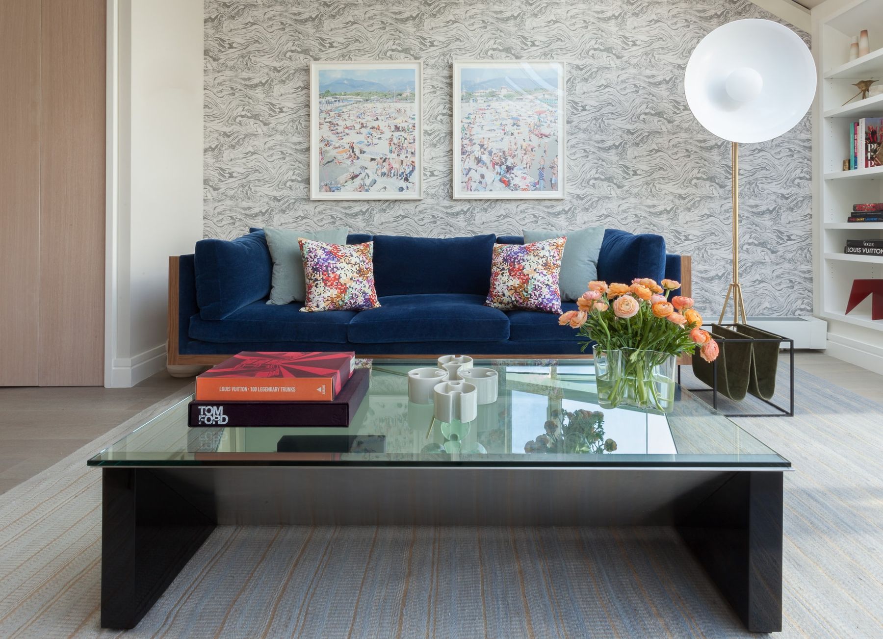 Study/Media room with blue velvet sofa in a walnut frame sits in front a gray and white abstract wallpaper, large glass coffee table with fashion books atop and large, Hollywood-style floor lamp