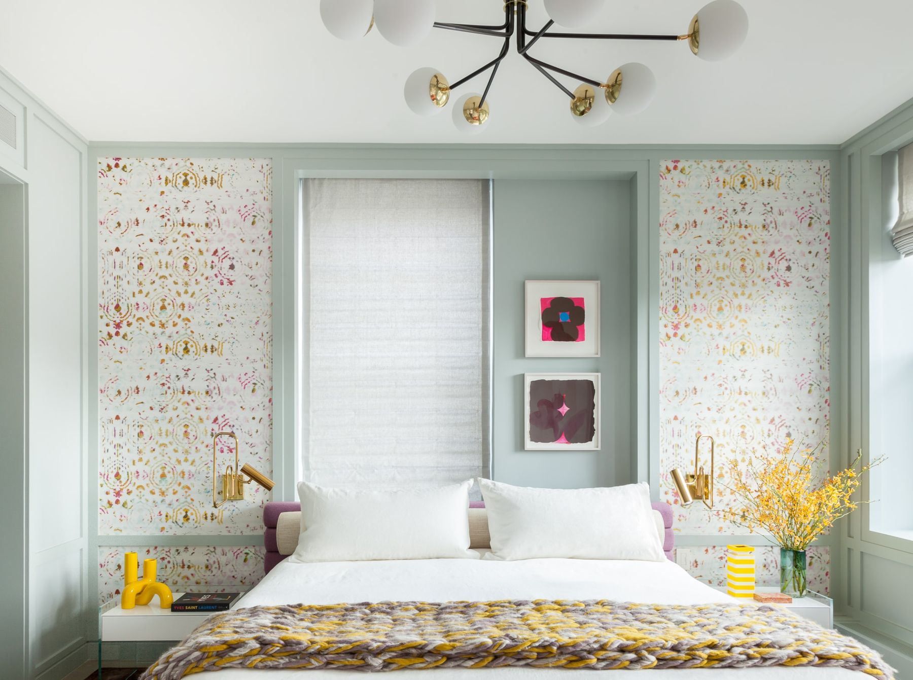 Primary bedroom with soft colors and patterned accent wallpaper, chunky quilt and matching white bedside tables and gold sconces