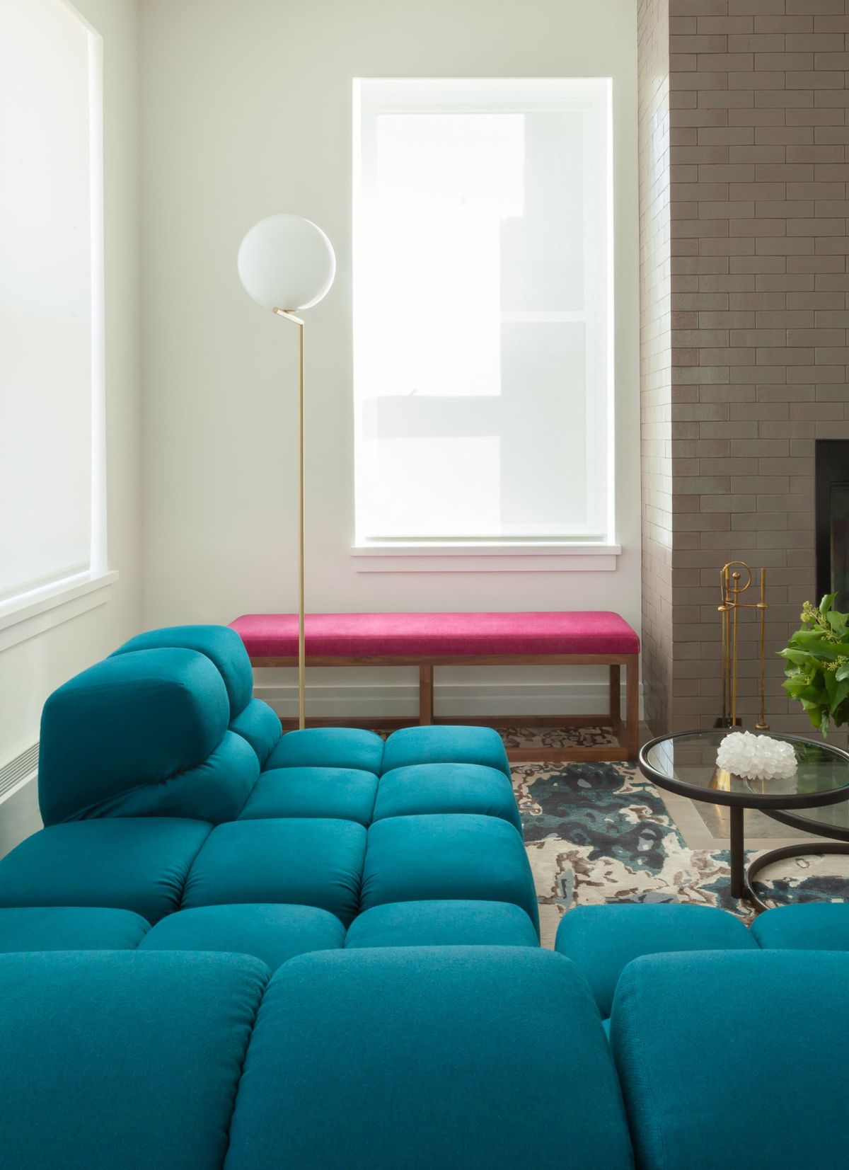 Vignette of teal sofa with magenta-cushioned bench nested beneath large window and adjacent to fireplace