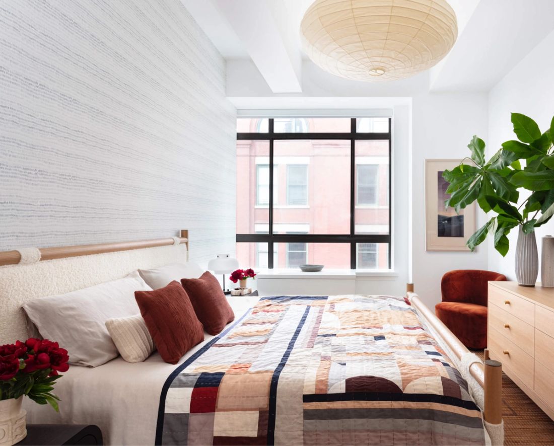 Primary bedroom with soft gray and white patterned wallpaper, boucle and wood bed frame, rust-colored velvet chair and wood 6-drawer dresser