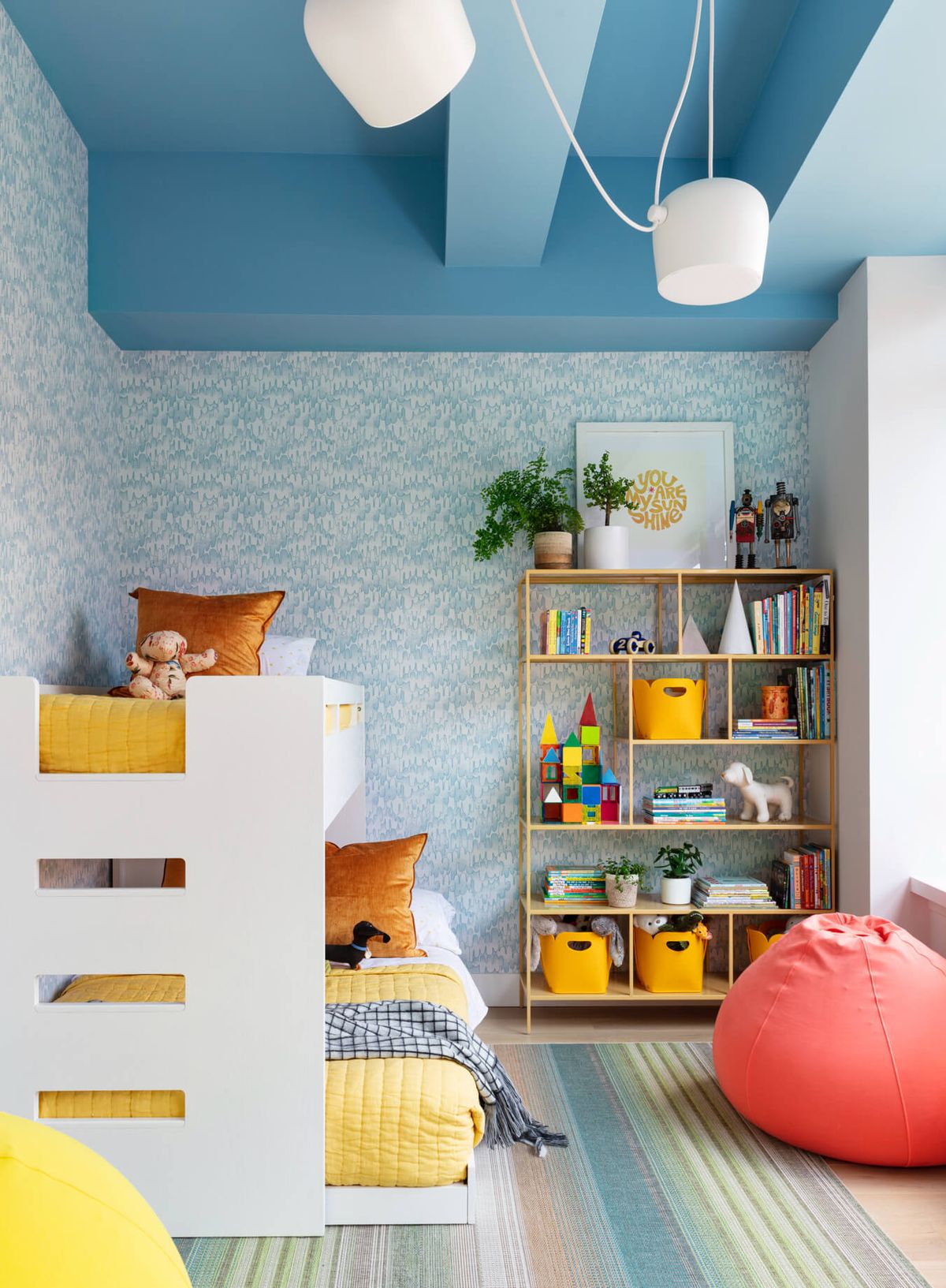 Child's room with white bunk beds, bookshelf, multi-colored rectangular rug, coral bean bag and blue and white patterned wallpaper 