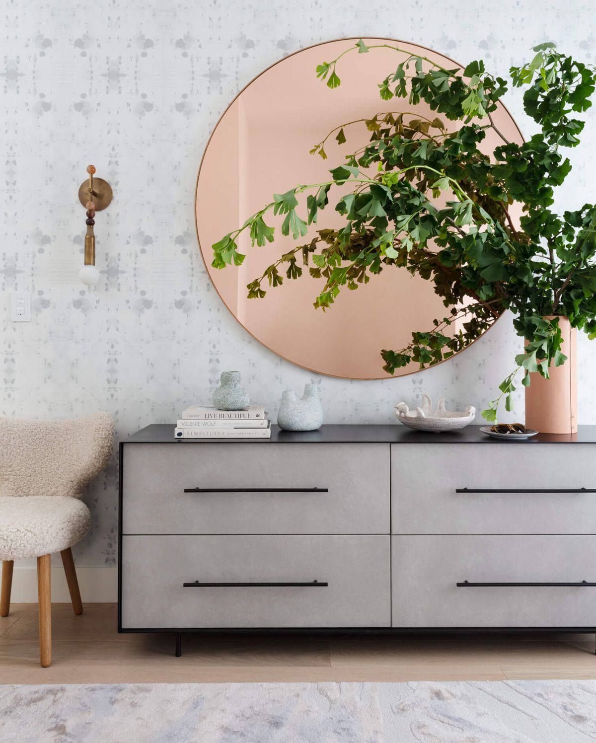 Foyer with large gray-toned 4-drawer dresser beneath very large, round, peach mirror with a lightly patterned gray and white wallpaper