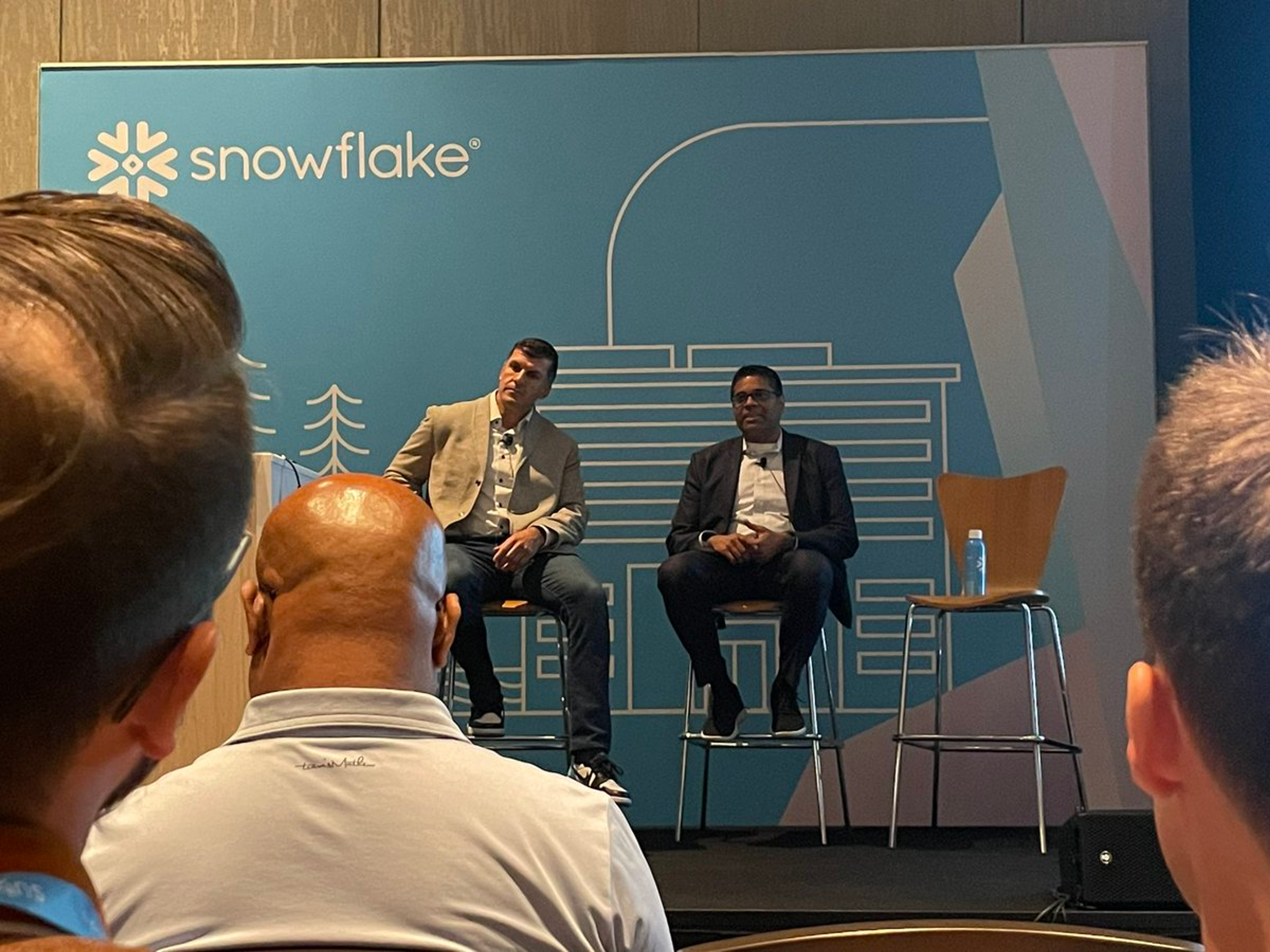 Two men sitting on stools in front of a blue sign that says Snowflake.