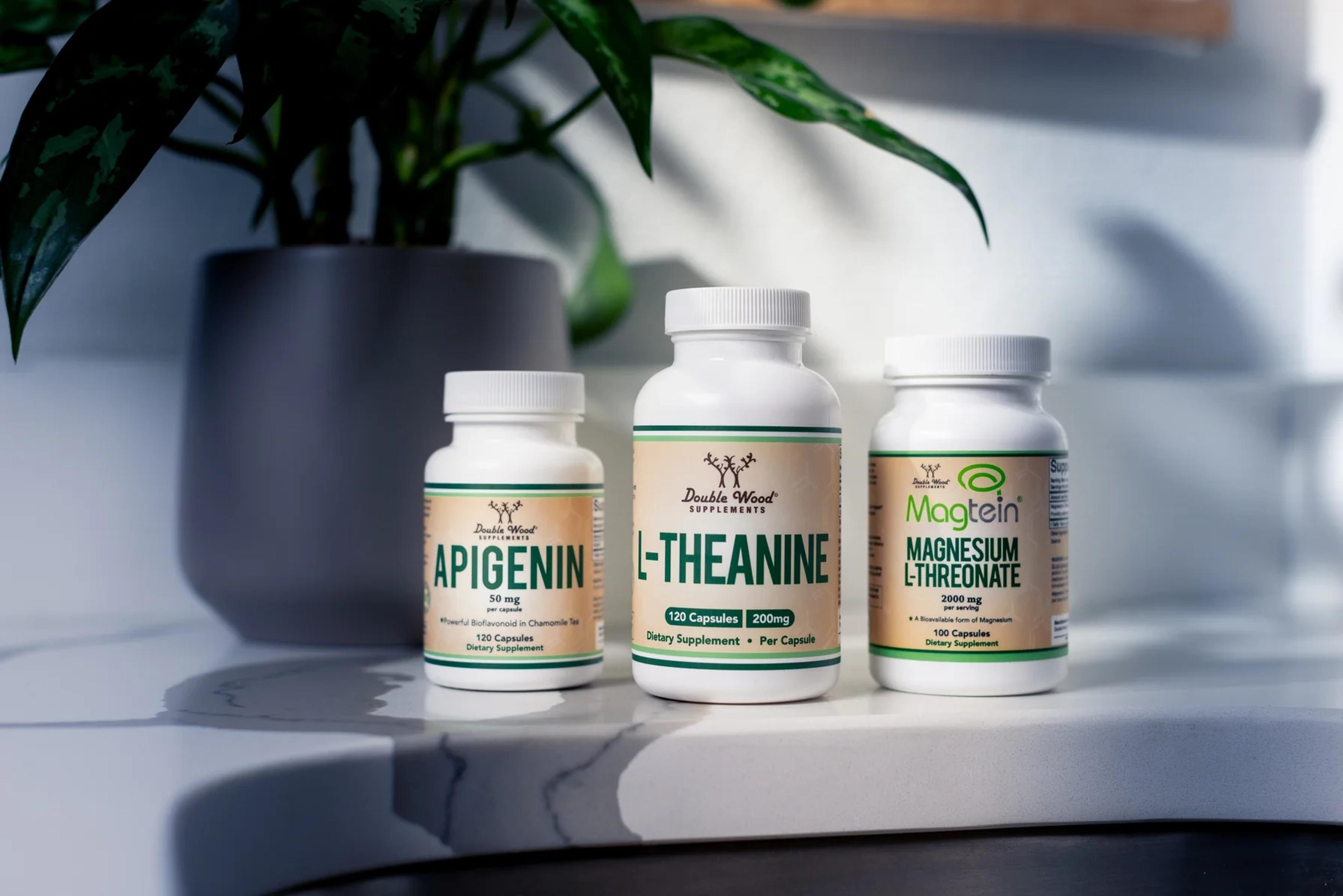 Three Double Wood Supplements bottles, Apigenin, L-Theanine and Magnesium L-Threonate, on a marble counter beside a potted plant