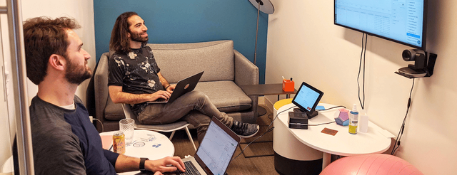 Two men sitting in front of a large screen with a laptop and a bottle of water.
