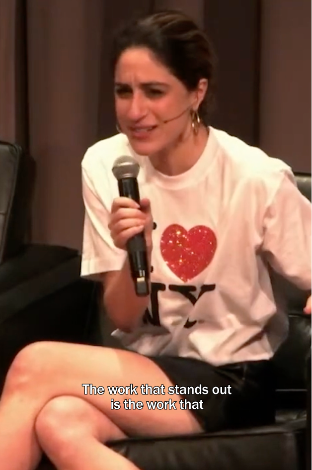 Talia Cotton speaks into a handheld microphone at Typographics, seated onstage and wearing a white T-shirt with a glittery red heart graphic. Subtitles read, “The work that stands out is the work that.