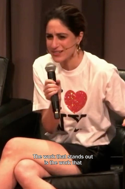 Talia Cotton speaks into a handheld microphone at Typographics, seated onstage and wearing a white T-shirt with a glittery red heart graphic. Subtitles read, “The work that stands out is the work that.