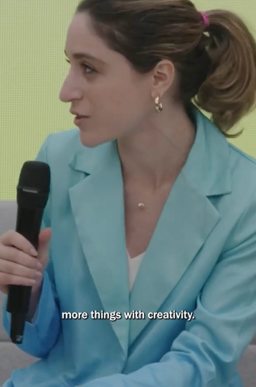 Talia Cotton sits in a light blue blazer, holding a black handheld microphone and speaking while looking to the side. Her brown hair is pulled back in a ponytail, and the on-screen caption reads, “more things with creativity.”