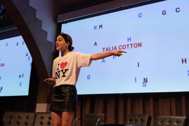 Talia Cotton stands on a stage and points toward a large presentation screen while speaking. She wears a white “I ♥ NY” T-shirt and black skirt. The screen displays scattered letters and the name “TALIA COTTON” in red, suggesting an interactive presentation or word game.