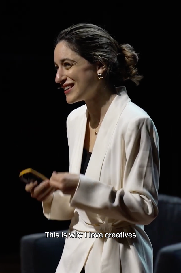 Talia Cotton stands on a dark stage, smiling as she speaks into a headset microphone while holding a smartphone. She wears a white blazer, her hair is tied back, and the caption reads, “This is why I love creatives.”