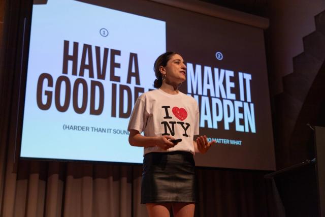 Talia Cotton stands on a stage in front of a large presentation screen, holding a presentation clicker and gesturing as she speaks. She wears a white “I ♥ NY” T-shirt and black skirt. The screen behind her displays large text reading “HAVE A GOOD IDEA MAKE IT HAPPEN.”