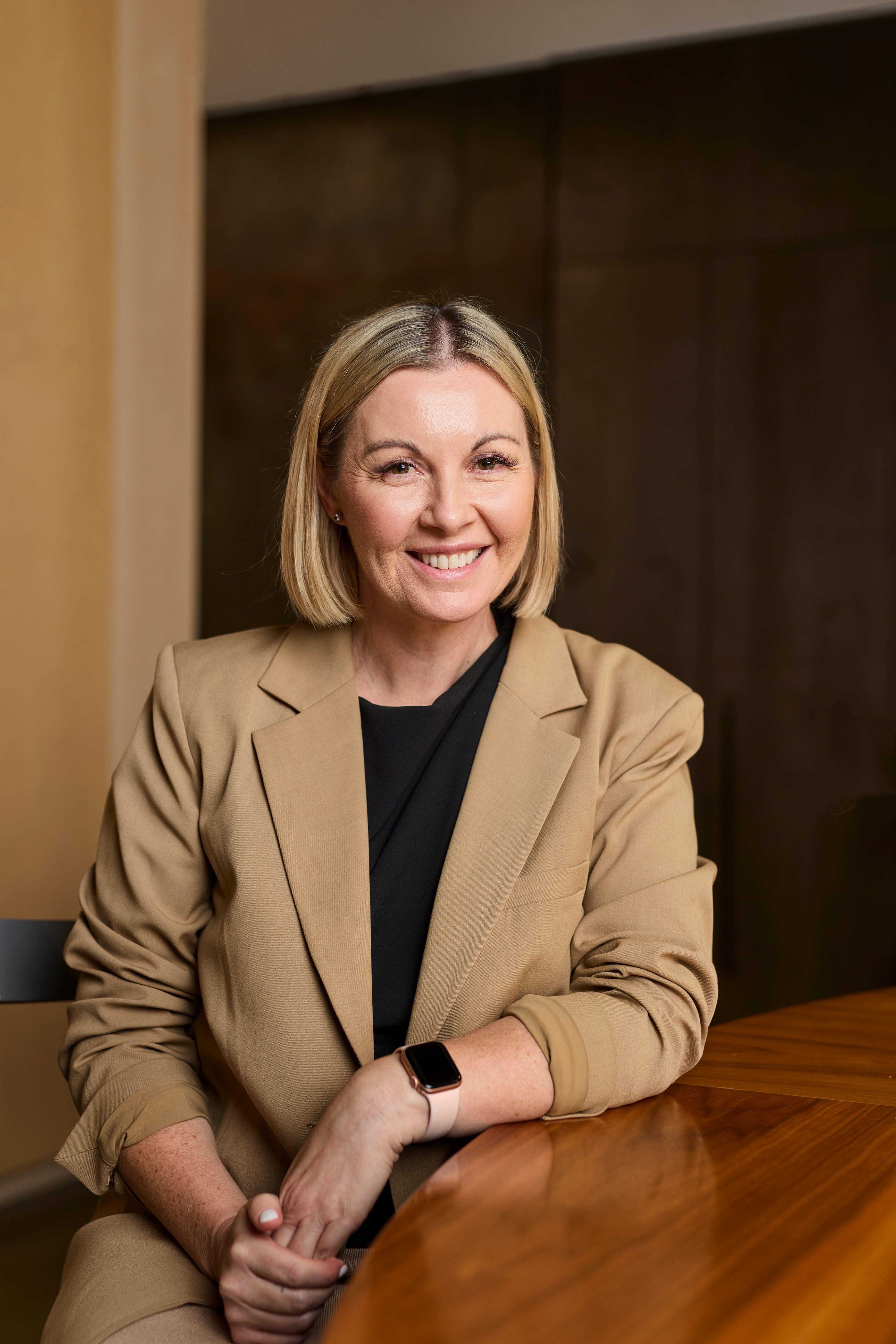 A woman in a beige blazer sits smiling to camera