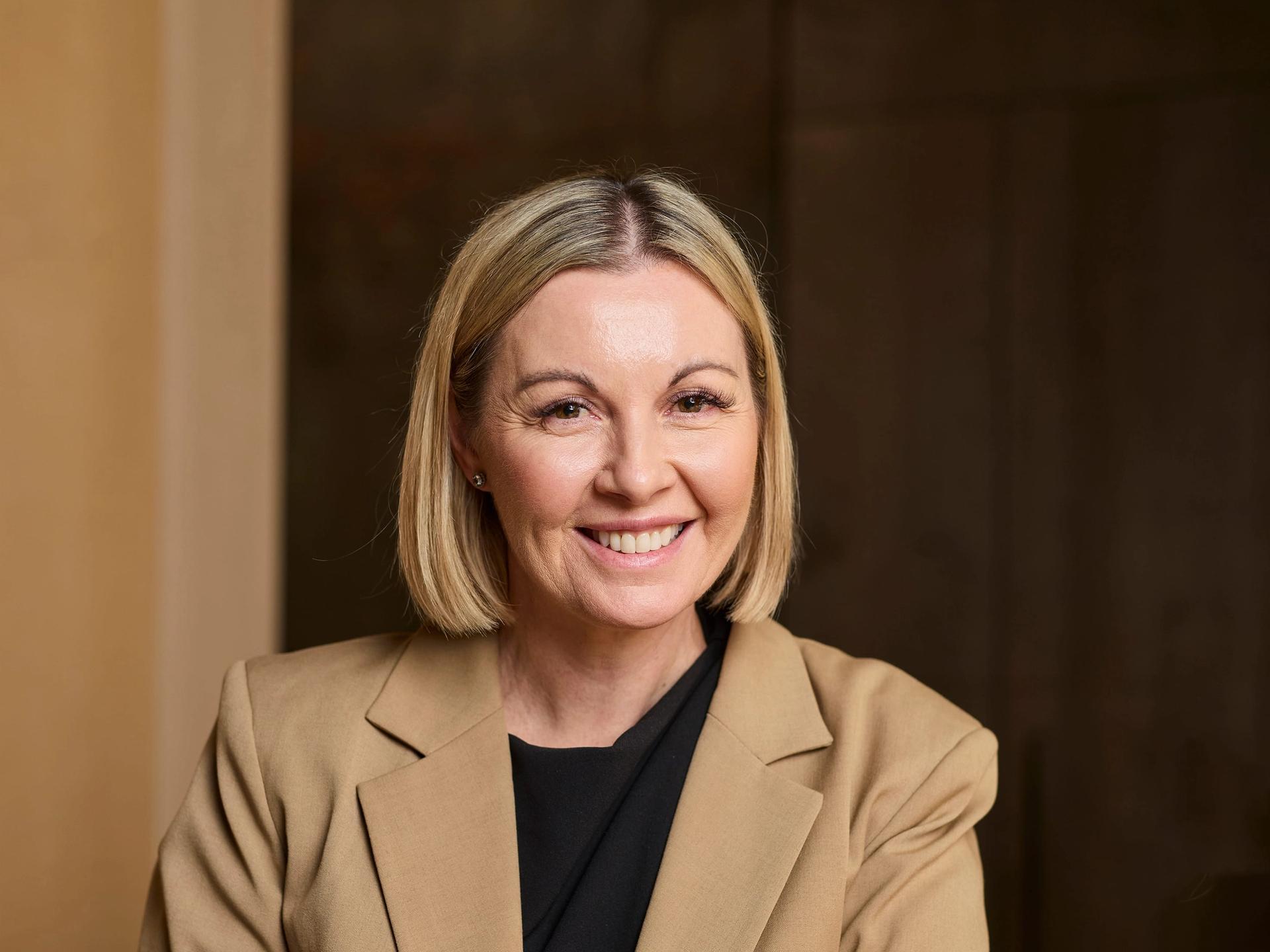 A woman in a beige blazer sits smiling to camera