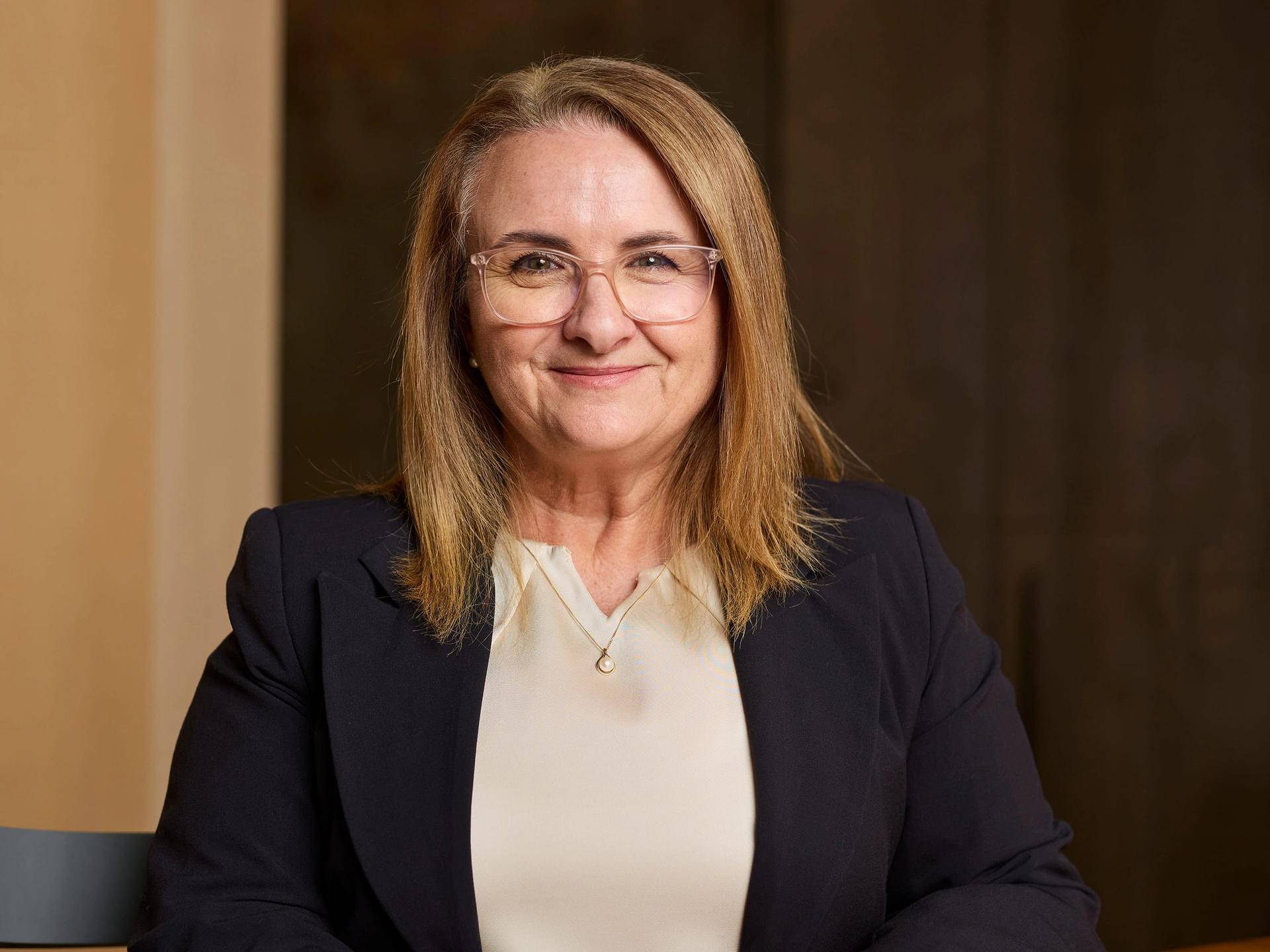 A woman in a blazer sits smiling to camera
